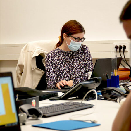 There are two students visible in this picture. They sit on opposite sides of a big office table. They both wear face masks. Each person is working on a laptop.